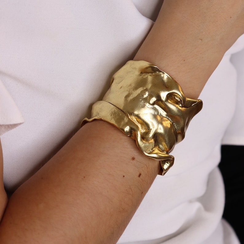 May include: A gold-colored cuff bracelet with a textured, organic design. The bracelet appears to be made of a malleable metal, with a rippled, flowing appearance. The cuff is worn on the arm, against a white background.