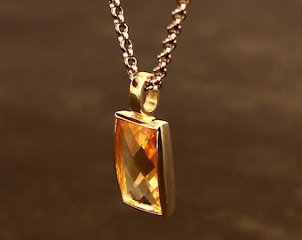 May include: A gold necklace featuring a rectangular, faceted, orange-yellow gemstone pendant. The pendant is set in a gold frame and suspended from a delicate chain. The background is a blurred, warm brown.