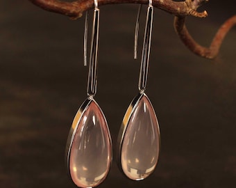 May include: A pair of drop earrings with a refined design. Each earring has a teardrop-shaped, pale pink gemstone set in a silver-toned metal frame. The earrings hang from long, slender silver hooks, creating a delicate look. The earrings are suspended from a brown branch.