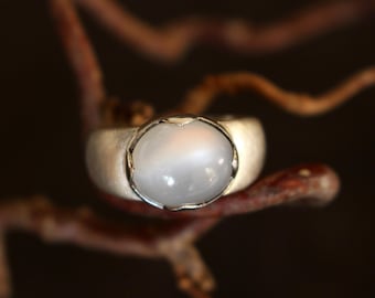May include: A silver ring with a large, oval, milky white gemstone. The ring has a brushed metal finish and a bezel setting. The gemstone is the focal point, set against a dark background, highlighting its smooth, polished surface.