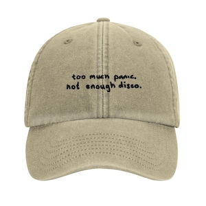 May include: A khaki-colored baseball cap with the embroidered text "too much panic. not enough disco." The cap has a curved brim and a six-panel design. The text is in a handwritten style.