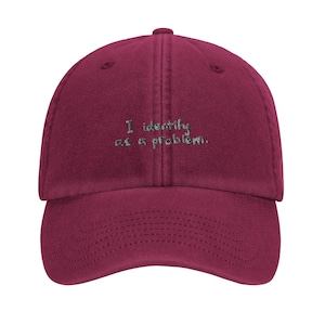 May include: A burgundy baseball cap with a curved peak. The cap has the embroidered text "I identify as a problem" in light green thread. The cap has a structured, six-panel design with a top button and adjustable strap.