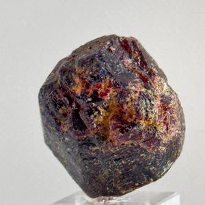 Puede incluir: Una gema de granate marrón rojizo oscuro con una estructura cristalina rugosa. La gema se exhibe sobre un soporte acrílico transparente sobre un fondo blanco. La piedra tiene una superficie texturizada con toques dorados y rojos.