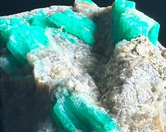 Può includere: Un gruppo di cristalli di colore verde smeraldo incastonati in una matrice rocciosa grigia e ruvida. I cristalli sono affilati e appuntiti, con un aspetto traslucido e vitreo.