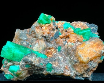 May include: A cluster of emerald green crystals embedded in a rough, brown, and gray rock matrix. The crystals are sharp and have a glassy luster.