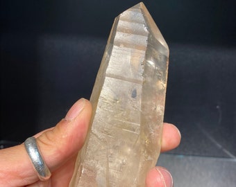 May include: A clear, light-brown quartz crystal with a pointed tip. The crystal is held in a hand, showcasing its natural, textured surface and unique shape. The crystal's color ranges from pale yellow to a light amber.
