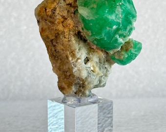 Könnte beinhalten: Ein roher, leuchtend grüner Smaragdkristall, eingebettet in eine texturierte, beige-braune Mineralmatrix. Das Exemplar ist sicher auf einem klaren, transparenten Acrylwürfel montiert, der den Kontrast zwischen Stein und Gestein betont.