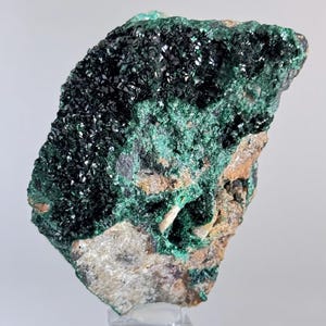 May include: A green and black malachite crystal specimen on a clear acrylic stand. The crystal has a rough, textured surface and is a beautiful example of this mineral.