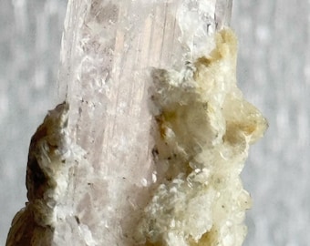May include: A large, clear, translucent crystal with a pink hue. The crystal is partially embedded in a white, opaque, rough matrix. The crystal is on a clear acrylic display stand.