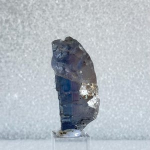 Op de afbeelding: Een helder kristal met een blauwe en paarse tint, getoond op een heldere acrylstandaard.