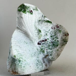 May include: A white, green, and red carved stone sculpture with a unique, organic shape. The sculpture is displayed on a clear acrylic stand.