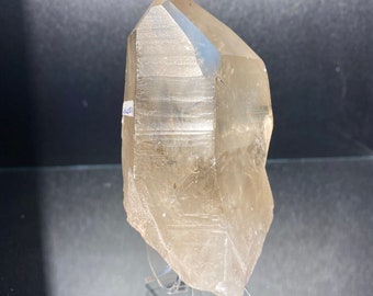May include: A large, translucent quartz crystal with a pointed tip and a rough, textured base. The crystal is a light amber color and is displayed on a clear stand against a dark background. The crystal is approximately 6 inches tall.