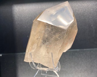 May include: A translucent, light brown quartz crystal with a faceted top and a rough, uneven base. The crystal is displayed on a clear acrylic stand against a dark background, highlighting its natural form and clarity.