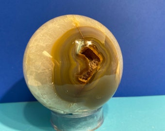 May include: A polished agate sphere, showcasing concentric bands of cream, brown, and gray. The crystal ball is mounted on a clear, circular stand. The center reveals a cavity with a dark brown interior, adding a unique geological feature.