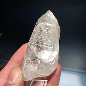 Op de afbeelding: Een helder kwartskristal met een puntige punt. Het kristal wordt ondersteund door een heldere, rechthoekige standaard. Het kristal heeft interne lijnen en een enigszins troebel uiterlijk. De achtergrond is donker.