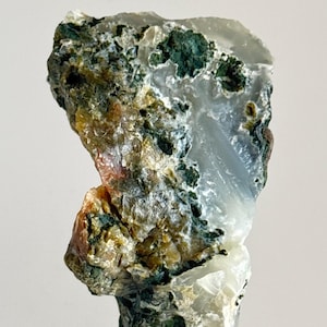 Puede incluir: Un espécimen de piedra preciosa natural en bruto. La piedra presenta una mezcla de colores, incluyendo blanco translúcido, verde intenso y toques de marrón y amarillo. La textura rugosa y la forma única sugieren que es un espécimen mineral, posiblemente para coleccionistas o joyería.