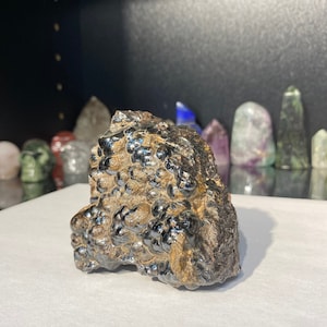 Könnte beinhalten: Ein großer, dunkelgrauer und brauner Hämatitkristall mit einem metallischen Glanz. Der Kristall hat eine raue, unregelmäßige Oberfläche mit vielen kleinen, abgerundeten Erhebungen.