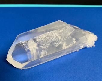 May include: A clear quartz crystal with a pointed tip and a rough, textured surface. The crystal is lying on a blue background.