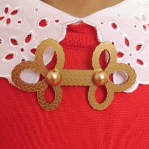 May include: A gold-colored, decorative, braided,  knot-shaped brooch with two round, gold-colored accents. The brooch is on a red sweater with a white, lacy collar.