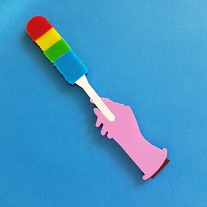May include: A pink hand holding a colorful cleaning brush with red, yellow, green, and blue bristles. The brush has a white handle.
