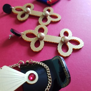 May include: A pair of gold-tone earrings with a floral design and pearl accents. The earrings are displayed on a red surface with a small blue and red drum pin.