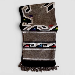 May include: A rectangular woven textile featuring a textured brown base with black, white, and grey geometric patterns. The design includes stepped motifs and small accents of red, blue, yellow, and green. The bottom edge has a brown fringe.