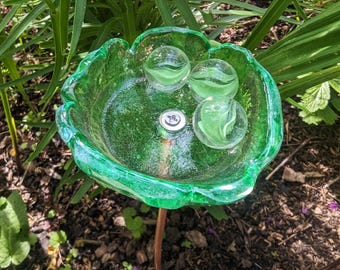 May include: A green glass flower-shaped garden stake featuring three clear glass marbles with white swirls inside the bowl. A silver metal screw is visible in the center of the glass bowl, which is attached to a thin copper-colored metal stem.