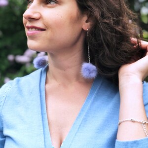 May include: A woman with long brown hair wearing a light blue cardigan with a deep V-neck. She is wearing a blue and white striped top underneath. She is wearing a pair of long, dangling earrings with purple pom-poms.