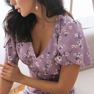 K&ouml;nnte beinhalten: Ein lila Kleid mit floralem Muster, V-Ausschnitt und kurzen, ausgestellten &Auml;rmeln. Das Kleid hat ein zartes Muster aus kleinen wei&szlig;en und roten Blumen. Das Model sitzt auf einem hellen Sofa.