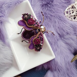 May include: A purple and gold beaded butterfly brooch with a large amethyst stone in the center. The brooch is on a white plate and a purple fur background.