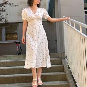 K&ouml;nnte beinhalten: Wei&szlig;es, knielanges Knopfkleid mit V-Ausschnitt und kurzen &Auml;rmeln. Das Kleid ist mit einem kleinen Blumenmuster in Lila- und Schwarzt&ouml;nen verziert. Das Model tr&auml;gt wei&szlig;e Sandalen.