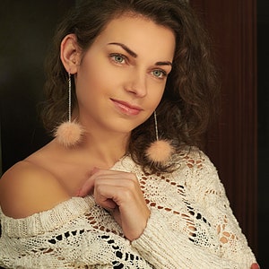 May include: A woman with long, curly brown hair wearing a white crocheted sweater and pink fluffy earrings.