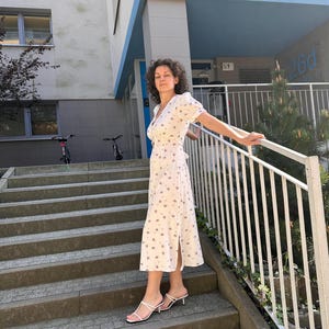 K&ouml;nnte beinhalten: Ein wei&szlig;es Wickelkleid mit floralem Muster und kurzen &Auml;rmeln. Das Kleid wird mit wei&szlig;en Riemchensandalen getragen. Die Frau steht auf Betontreppen mit einem wei&szlig;en Gel&auml;nder.