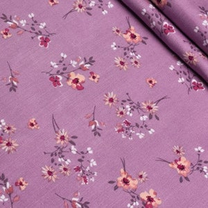 K&ouml;nnte beinhalten: Nahaufnahme eines lila Stoffes mit floralem Muster. Das Design zeigt kleine, zarte Bl&uuml;ten in Pfirsich-, Wei&szlig;- und Dunkelrosa-T&ouml;nen, die &uuml;ber die Oberfl&auml;che verstreut sind. Der Stoff ist gefaltet und zeigt seine Textur und den detaillierten Blumendruck.