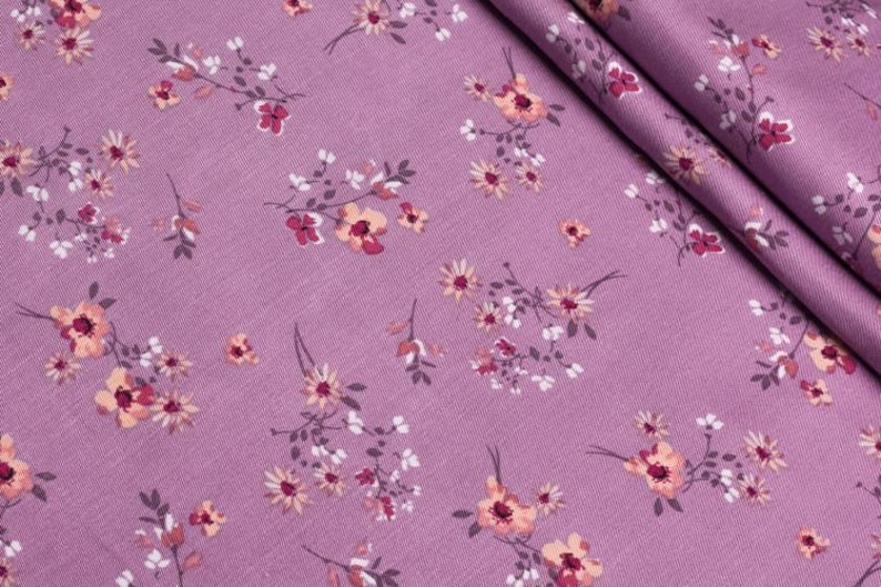 K&ouml;nnte beinhalten: Nahaufnahme eines lila Stoffes mit floralem Muster. Das Design zeigt kleine, zarte Bl&uuml;ten in Pfirsich-, Wei&szlig;- und Dunkelrosa-T&ouml;nen, die &uuml;ber die Oberfl&auml;che verstreut sind. Der Stoff ist gefaltet und zeigt seine Textur und den detaillierten Blumendruck.