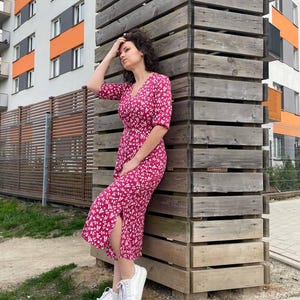 May include: A woman wearing a pink floral midi dress with a white background and white sneakers. She is standing next to a wooden structure.