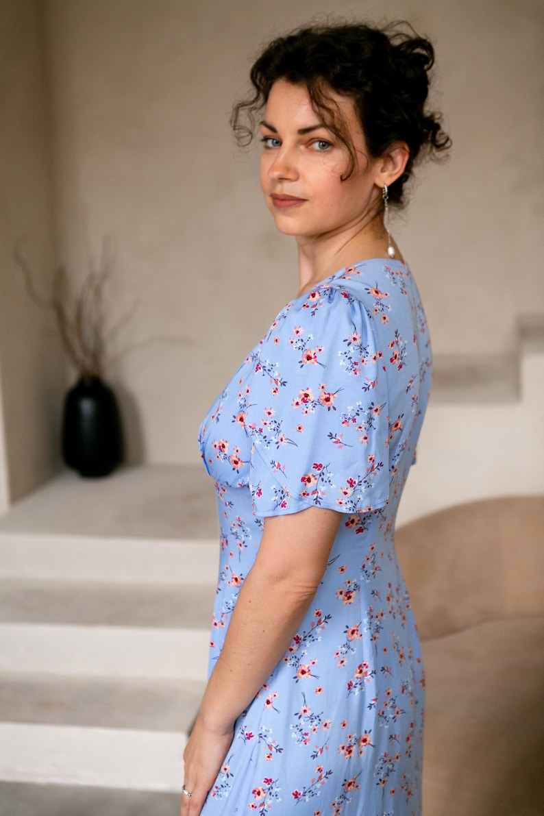 K&ouml;nnte beinhalten: Hellblaues Kleid mit Blumenmuster und kurzen &Auml;rmeln. Das Kleid hat einen V-Ausschnitt und ein Muster aus kleinen rosa und roten Blumen. Das Kleid ist aus leichtem Stoff gefertigt, ideal f&uuml;r warmes Wetter. Die Person tr&auml;gt Perlenohrringe.