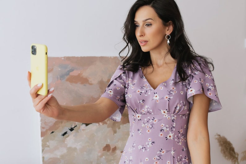 K&ouml;nnte beinhalten: Eine Person in einem lavendelfarbenen Kleid mit Blumenmuster und kurzen &Auml;rmeln h&auml;lt ein gelbes Smartphone. Das Kleid hat einen V-Ausschnitt und ein figurbetontes Oberteil. Der Hintergrund umfasst ein Gem&auml;lde mit neutralen Farben.