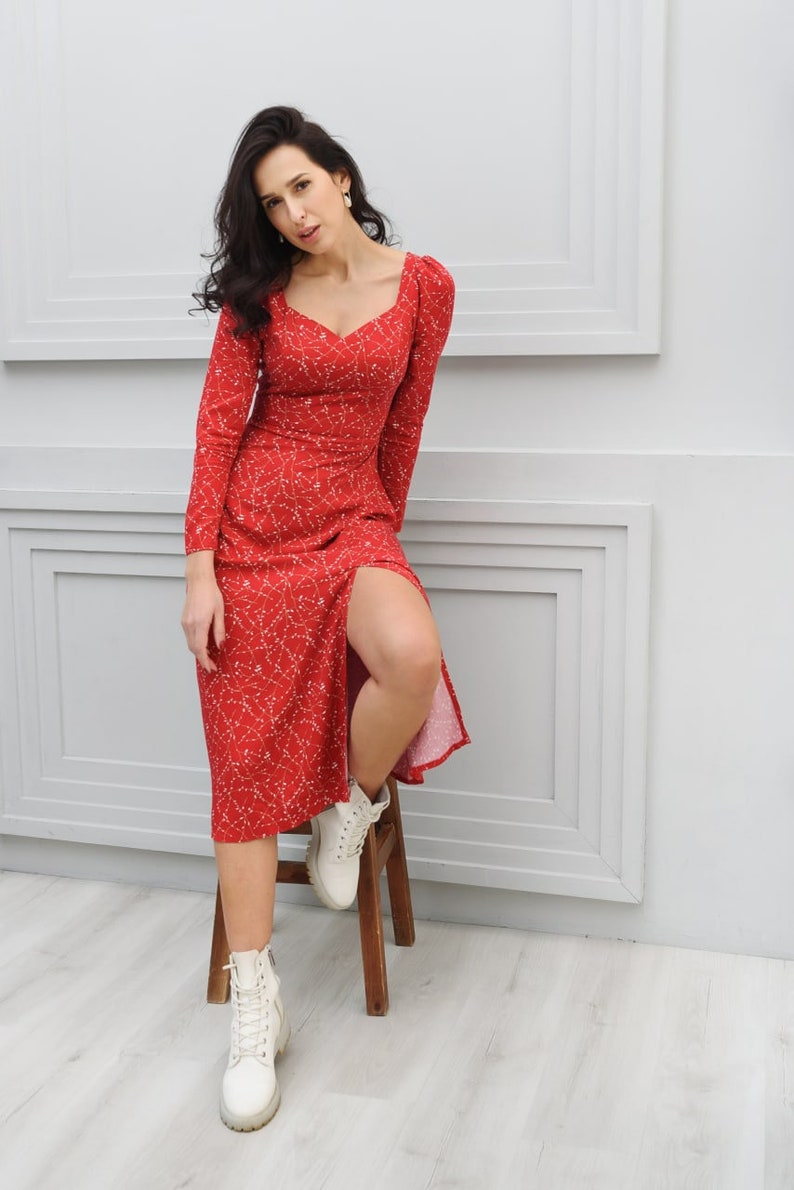 May include: A woman wearing a red midi dress with a white floral pattern and white lace-up boots. She is sitting on a wooden stool in front of a white wall.