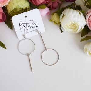 Puede incluir: Pendientes asimétricos plateados. Uno presenta un círculo grande con una barra vertical, el otro un círculo más pequeño con una barra. Los pendientes están sobre una superficie blanca con flores rosas, blancas y verdes.