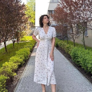 K&ouml;nnte beinhalten: Ein wei&szlig;es Kleid mit floralem Muster, kurzen &Auml;rmeln und einer Knopfleiste vorne. Das knielange Kleid wird mit wei&szlig;en Sandalen getragen. Der Hintergrund zeigt B&auml;ume und ein Geb&auml;ude.