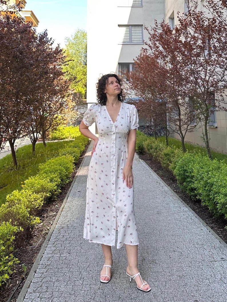 K&ouml;nnte beinhalten: Ein wei&szlig;es Kleid mit floralem Muster, kurzen &Auml;rmeln und einer Knopfleiste vorne. Das knielange Kleid wird mit wei&szlig;en Sandalen getragen. Der Hintergrund zeigt B&auml;ume und ein Geb&auml;ude.