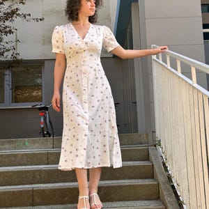 K&ouml;nnte beinhalten: Wei&szlig;es Midikleid mit floralem Muster und Knopfleiste vorne. Das Kleid hat kurze, gepuffte &Auml;rmel und einen V-Ausschnitt. Die Person tr&auml;gt wei&szlig;e Sandalen. Das Kleid ist etwa 114 cm lang.