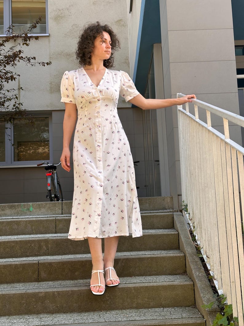 K&ouml;nnte beinhalten: Wei&szlig;es Midikleid mit floralem Muster und Knopfleiste vorne. Das Kleid hat kurze, gepuffte &Auml;rmel und einen V-Ausschnitt. Die Person tr&auml;gt wei&szlig;e Sandalen. Das Kleid ist etwa 114 cm lang.