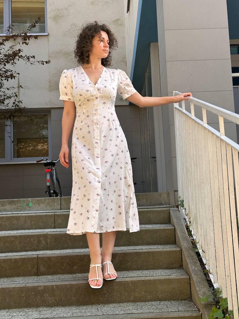 K&ouml;nnte beinhalten: Wei&szlig;es, knielanges Knopfkleid mit V-Ausschnitt und kurzen &Auml;rmeln. Das Kleid ist mit einem kleinen Blumenmuster in Lila- und Schwarzt&ouml;nen verziert. Das Model tr&auml;gt wei&szlig;e Sandalen.