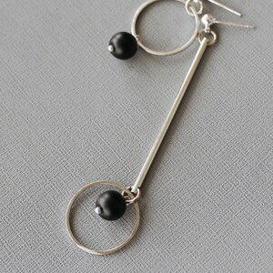 May include: A pair of silver earrings with black beads. The earrings are made of a thin silver rod with a circle at the bottom. The circle is attached to the rod with a black bead. The other earring is a simple circle with a black bead hanging from it.