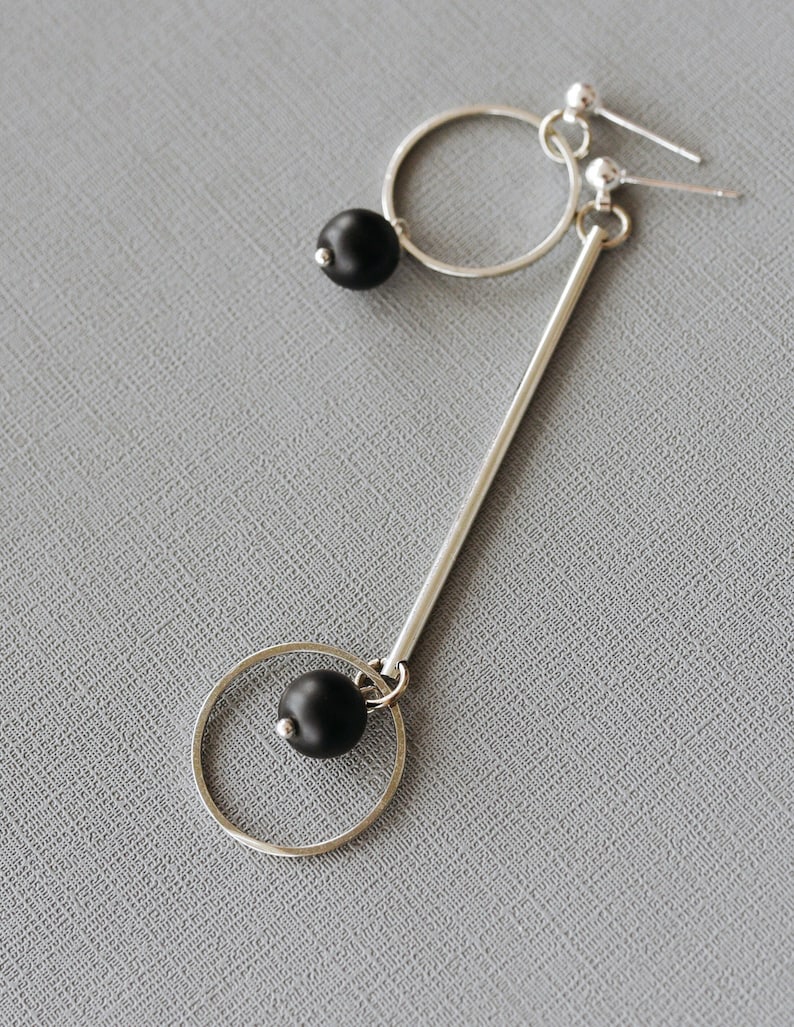 May include: A pair of silver earrings with black beads. The earrings are made of a thin silver rod with a circle at the bottom. The circle is attached to the rod with a black bead. The other earring is a simple circle with a black bead hanging from it.