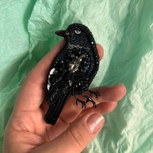 Peut inclure: Une broche en forme de corbeau noir faite de paillettes et de perles. Le corbeau a un œil en cristal et un bec détaillé.