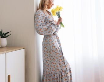 Puede incluir: Una mujer con un vestido maxi floral azul claro con un dobladillo con volantes se para junto a una ventana, sosteniendo un ramo de narcisos amarillos.