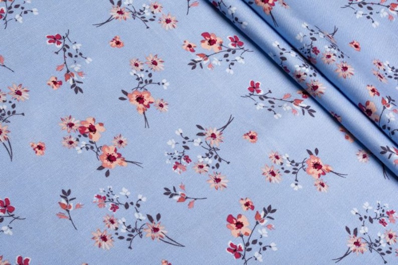 K&ouml;nnte beinhalten: Hellblauer Stoff mit einem sich wiederholenden Blumenmuster. Das Design zeigt kleine Gruppen von Bl&uuml;ten in Rosa, Pfirsich und Wei&szlig;, mit dunkelgr&uuml;nen Stielen und Bl&auml;ttern. Der Stoff ist gefaltet und zeigt seine Textur.